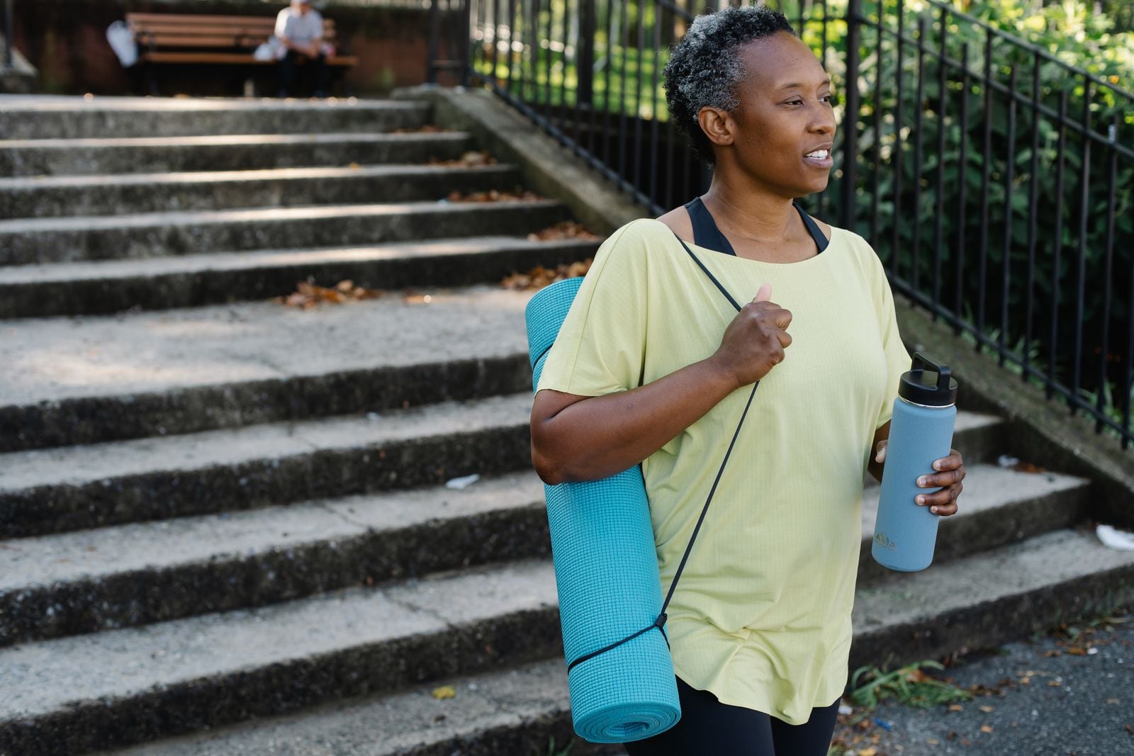 Smiling woman in workout clothes carrying a yoga mat and water bottle outdoors