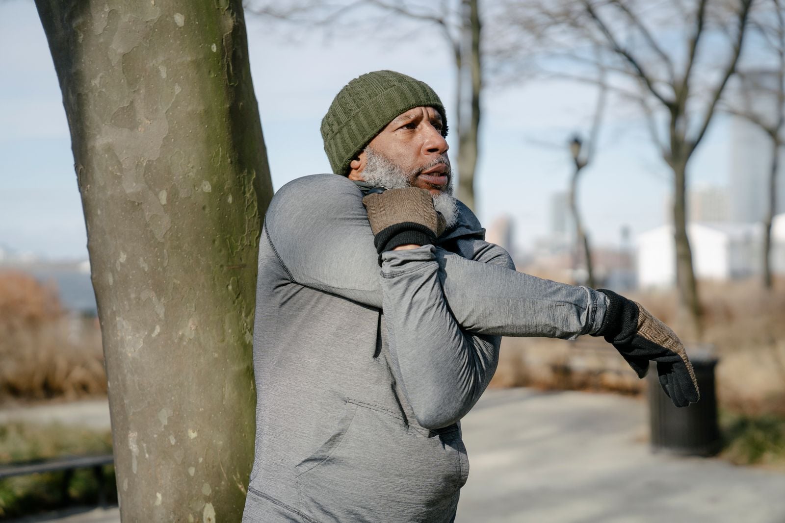 Older man doing a cross-body shoulder stretch beside a tree in a park