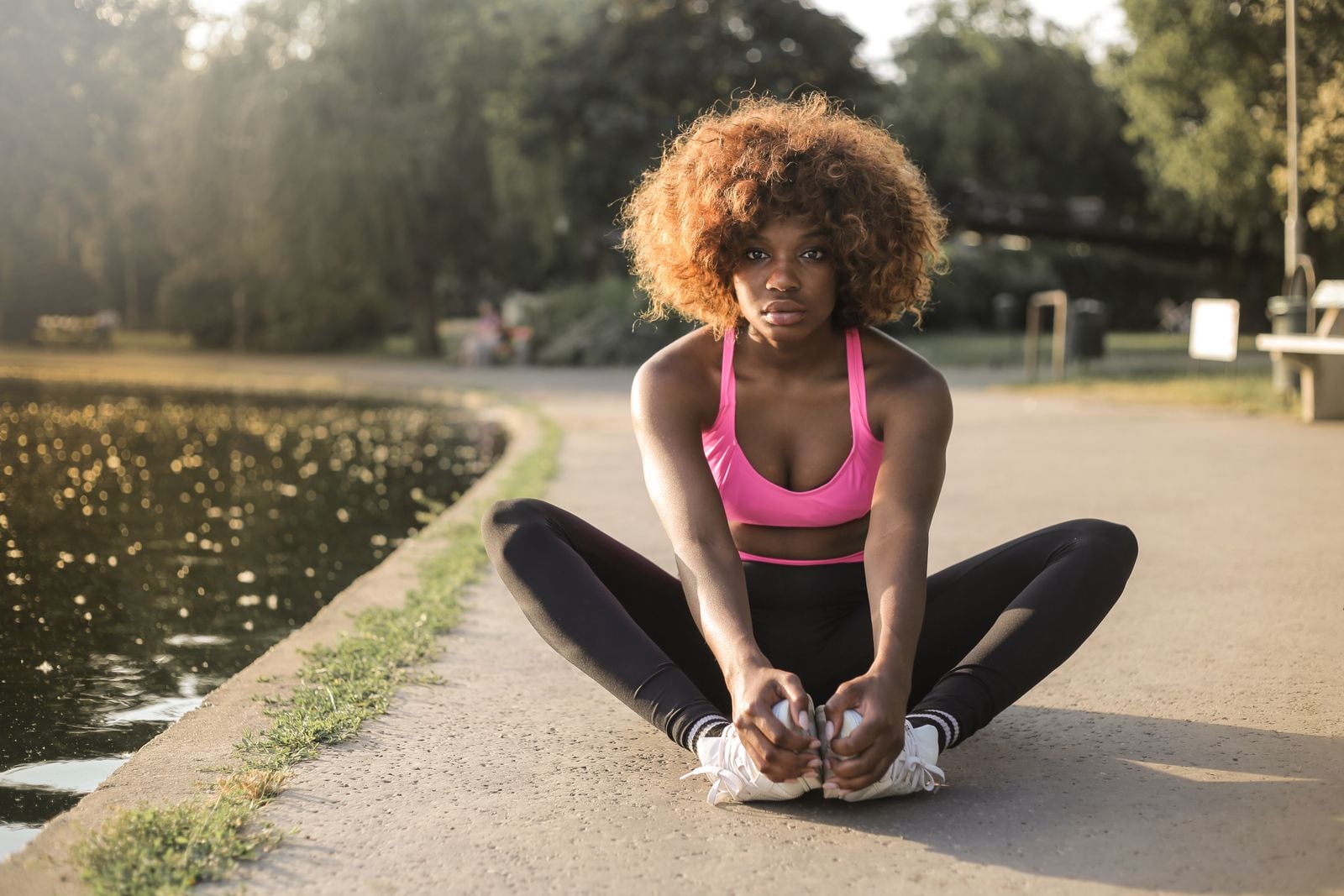 Woman doing a seated stretch on a park path beside a pond